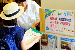 「夏休みユーラシア新聞を作ろう」と書かれた看板の前で、クリップボードを渡される麦わら帽子の少女。